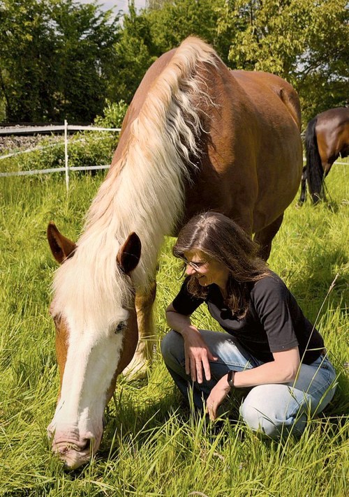 Eine Frau kniet im hohen Gras, ein helles Pferd senkt ihr den Kopf entgegen